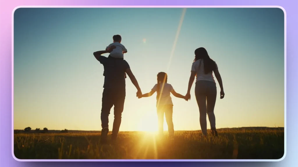Silhouetted family of four holding hands walking in a grassy field at sunset.