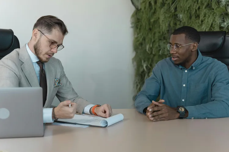 Two men in an office setting, one in a gray suit reviewing documents, the other in a blue shirt listening.