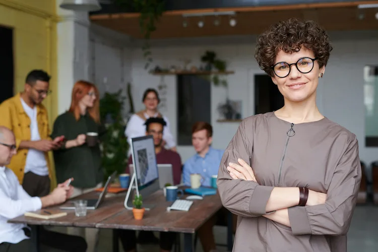 Young professional woman with short curly hair and glasses standing confidently in modern office with colleagues working in background.