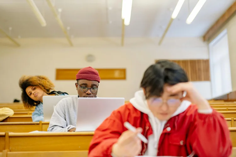 Three students seated in a classroom, two using laptops and one writing on paper.