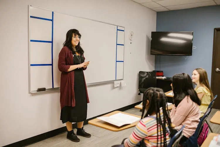 Female teacher standing by whiteboard instructing three attentive students in classroom setting.