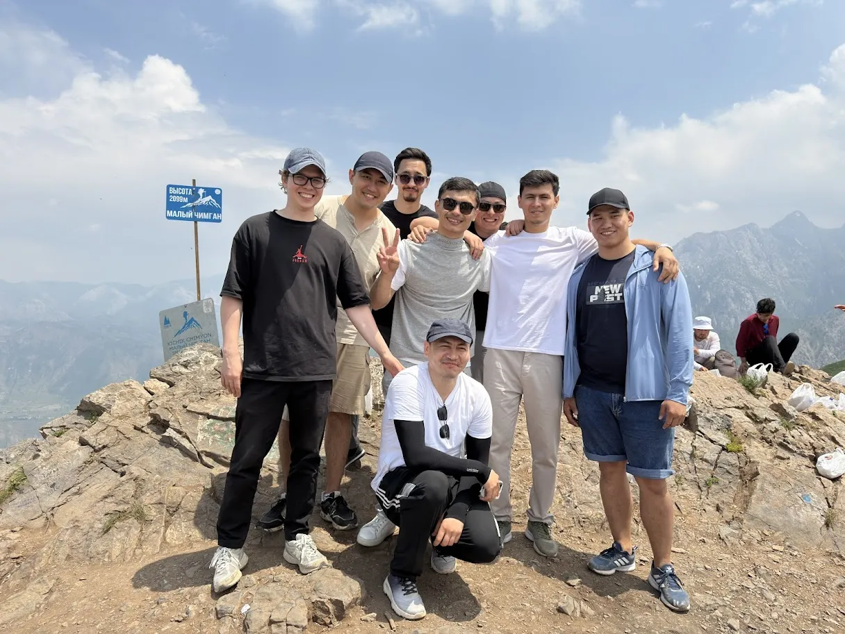 Group of eight young men posing on rocky mountain summit with blue sky and distant peaks.