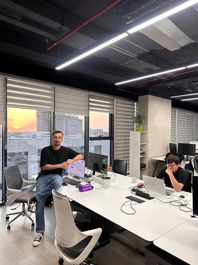 Modern office interior with two men working at desks, large windows, and sunset view.