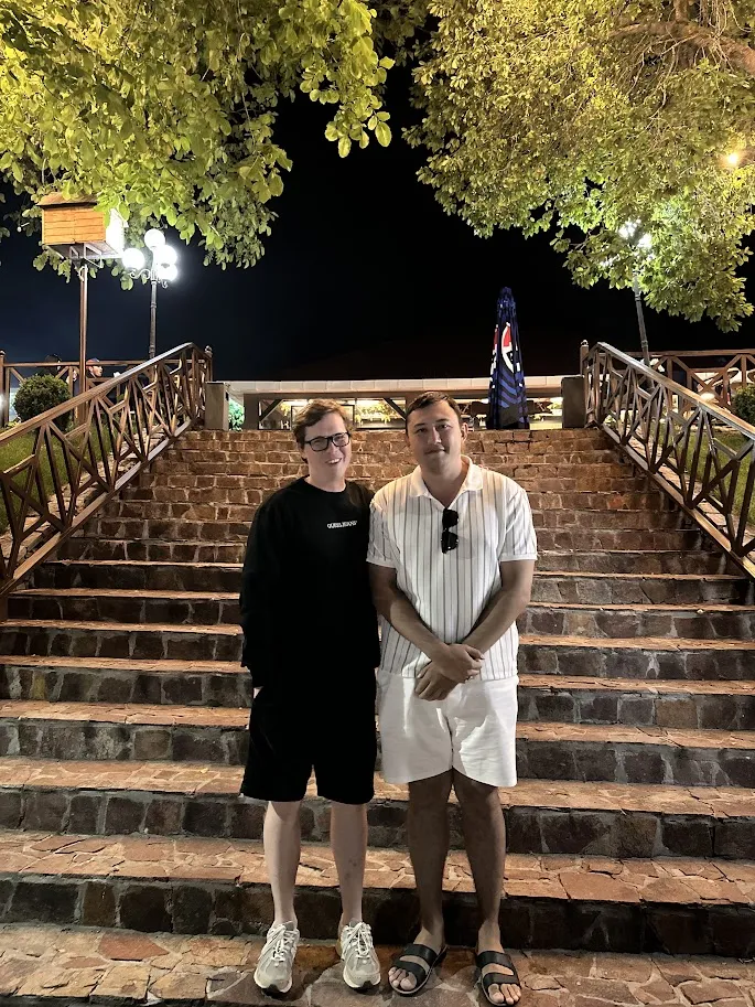 Two men standing on stone steps at night under illuminated green trees, with a dark sky background.