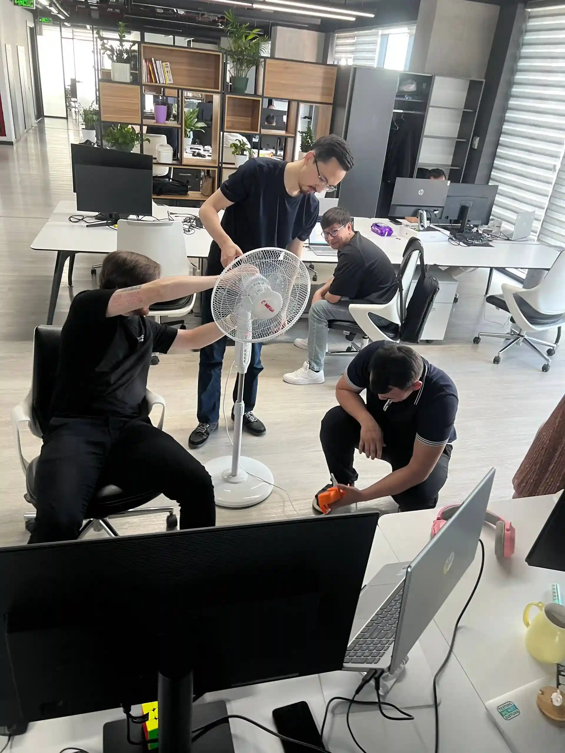 Office setting with four men assembling a white standing fan near desks and computers.