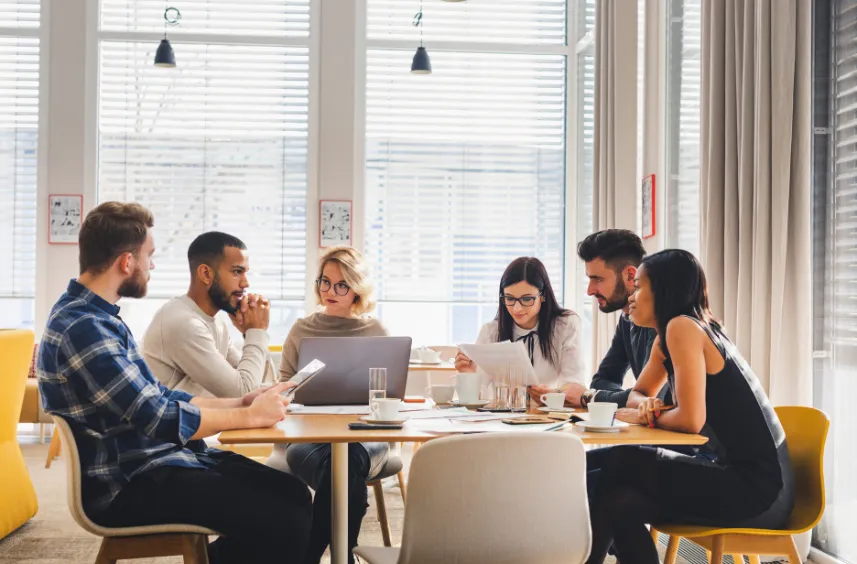 Six diverse professionals engaged in a meeting around a wooden table with laptops and documents.